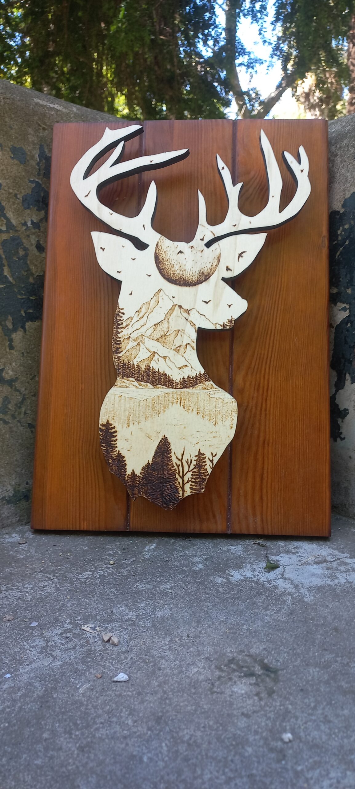 Silueta dun cervo con grandes cornos tallada en madeira, coa cabeza rechea dunha paisaxe detallada de montañas, árbores e paxaros. A obra está feita en tres táboas verticais de madeira pulida e aparece apoiada no exterior, sobre cemento, con árbores ao fondo.