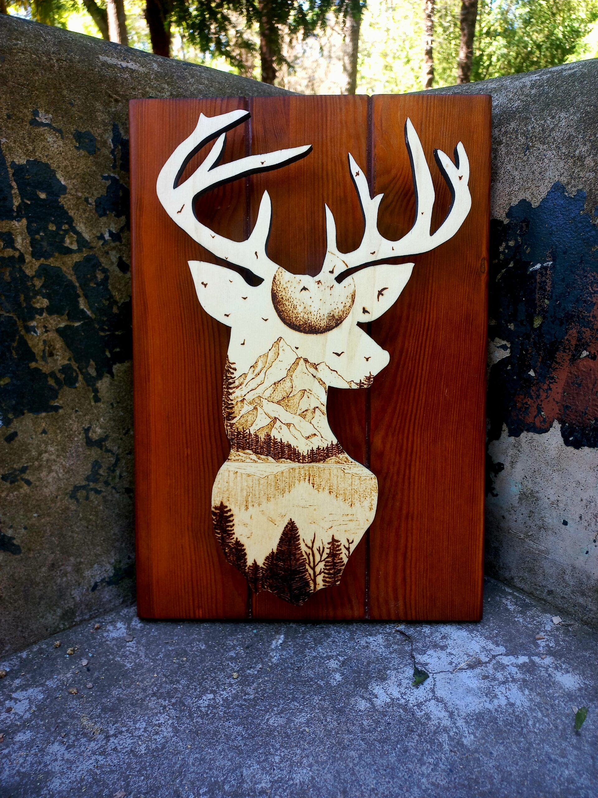 Silueta dun cervo con grandes cornos tallada en madeira, coa cabeza rechea dunha paisaxe detallada de montañas, árbores e paxaros. A obra está feita en tres táboas verticais de madeira pulida e aparece apoiada no exterior, sobre cemento, con árbores ao fondo.