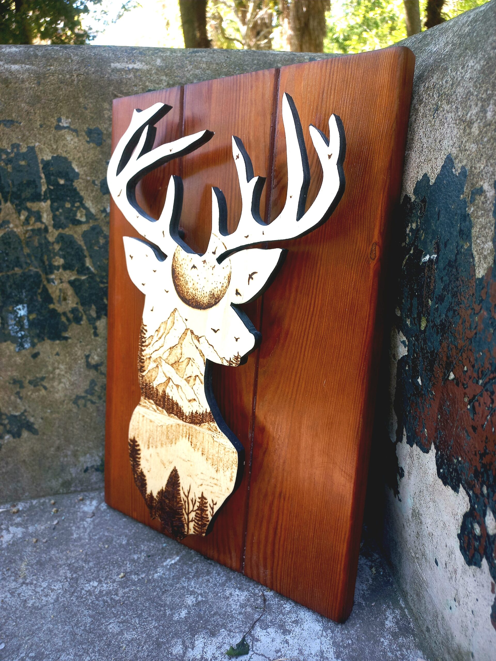 Silueta dun cervo con grandes cornos tallada en madeira, coa cabeza rechea dunha paisaxe detallada de montañas, árbores e paxaros. A obra está feita en tres táboas verticais de madeira pulida e aparece apoiada no exterior, sobre cemento, con árbores ao fondo.