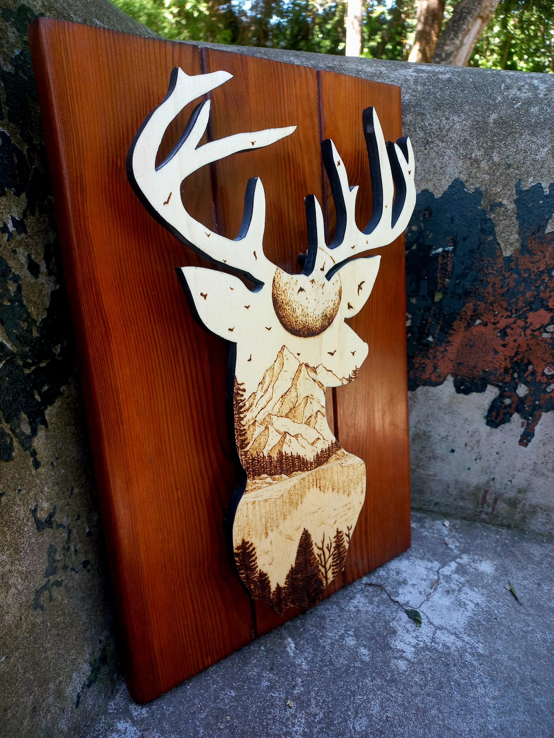 Silueta dun cervo con grandes cornos tallada en madeira, coa cabeza rechea dunha paisaxe detallada de montañas, árbores e paxaros. A obra está feita en tres táboas verticais de madeira pulida e aparece apoiada no exterior, sobre cemento, con árbores ao fondo.