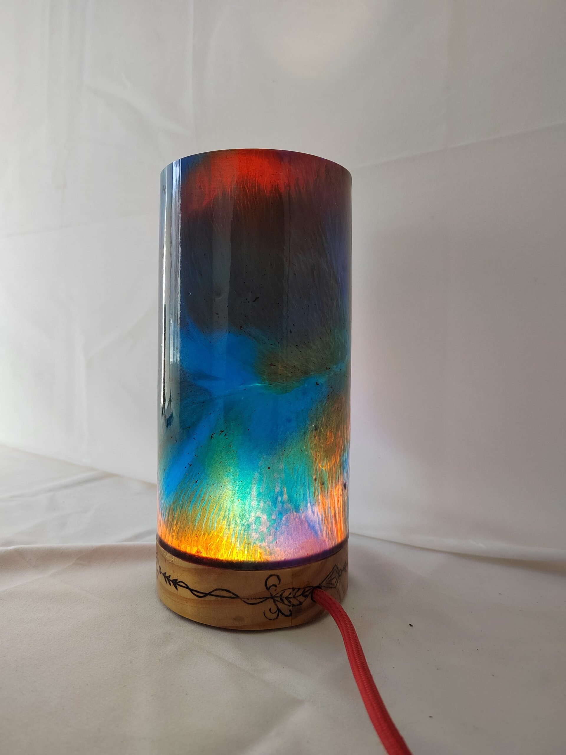Lámpada artesanal con corpo cilíndrico de resina en cores vibrantes e base de madeira gravada, iluminada desde o interior e unida cunha costura decorativa de fío metálico.