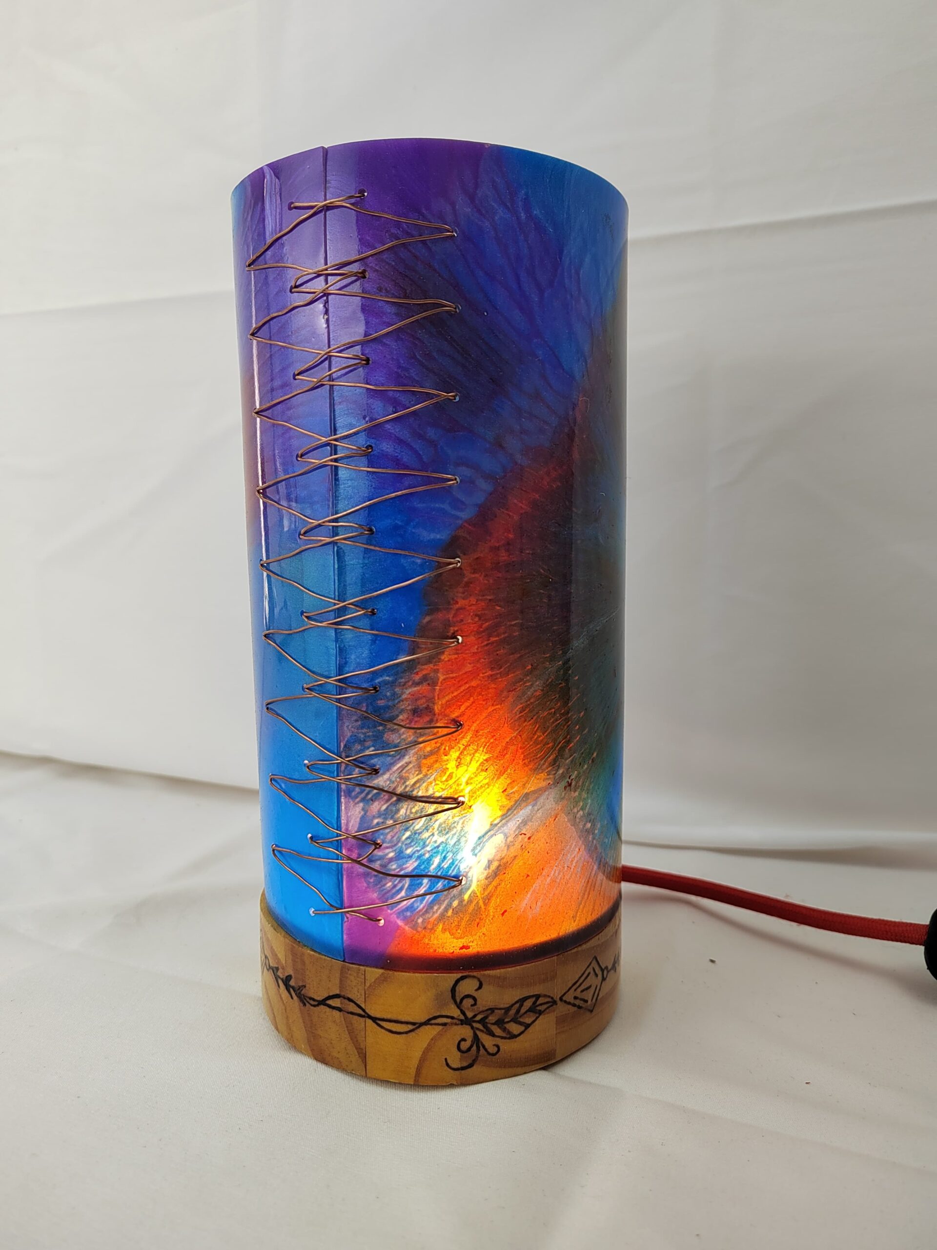 Lámpada artesanal con corpo cilíndrico de resina en cores vibrantes e base de madeira gravada, iluminada desde o interior e unida cunha costura decorativa de fío metálico.