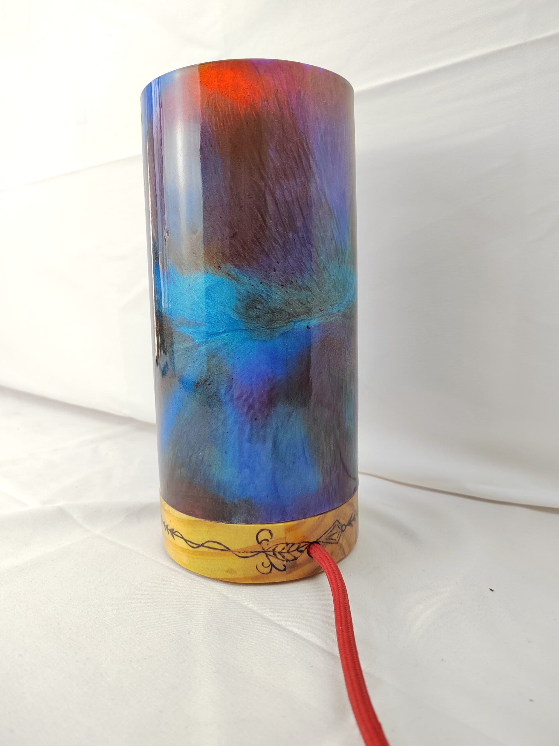 Lámpada artesanal con corpo cilíndrico de resina en cores vibrantes e base de madeira gravada, iluminada desde o interior e unida cunha costura decorativa de fío metálico.