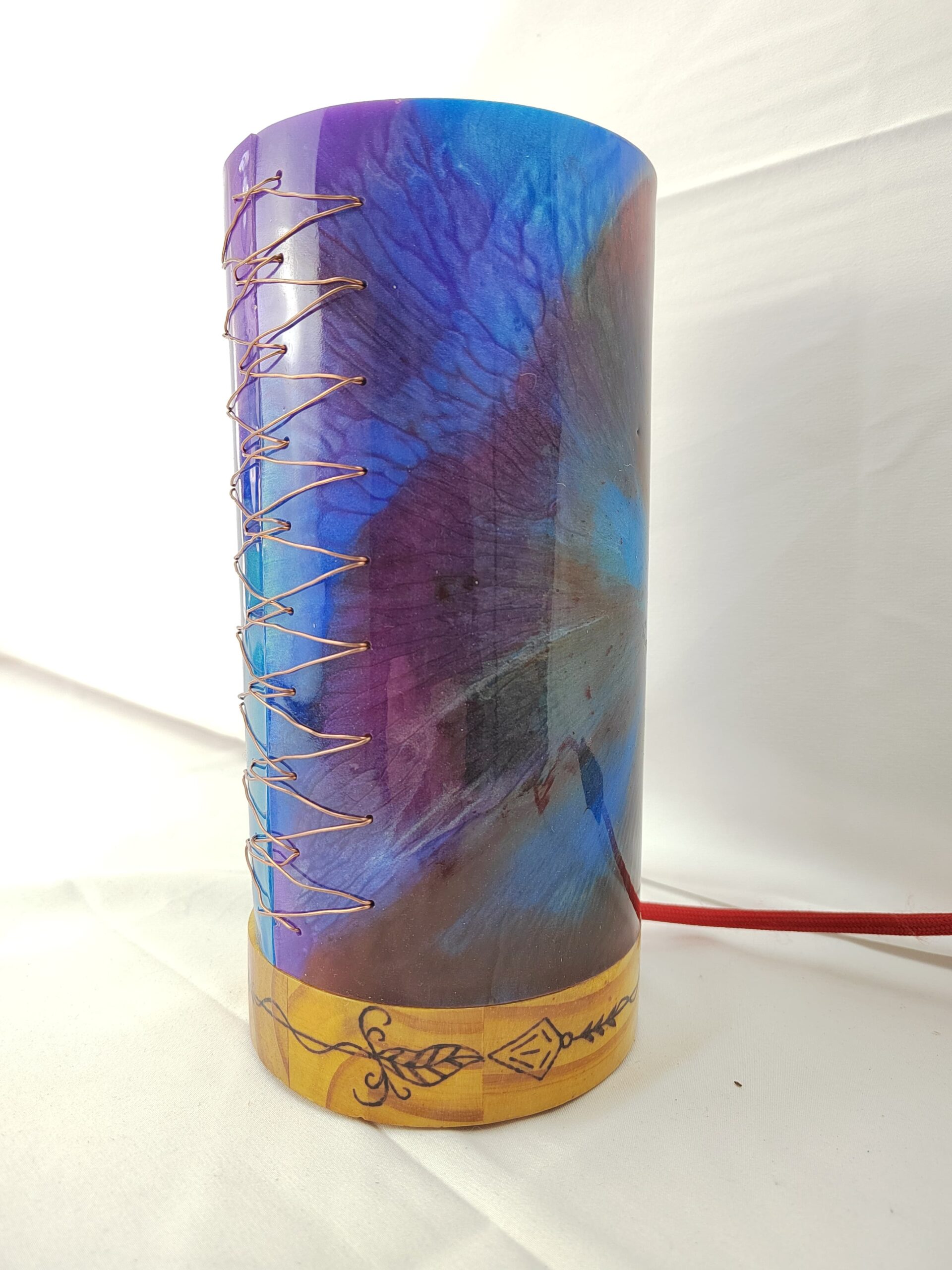 Lámpada artesanal con corpo cilíndrico de resina en cores vibrantes e base de madeira gravada, iluminada desde o interior e unida cunha costura decorativa de fío metálico.