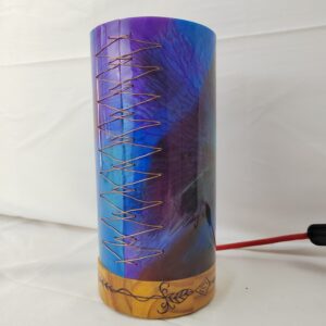 Lámpada artesanal con corpo cilíndrico de resina en cores vibrantes e base de madeira gravada, iluminada desde o interior e unida cunha costura decorativa de fío metálico.