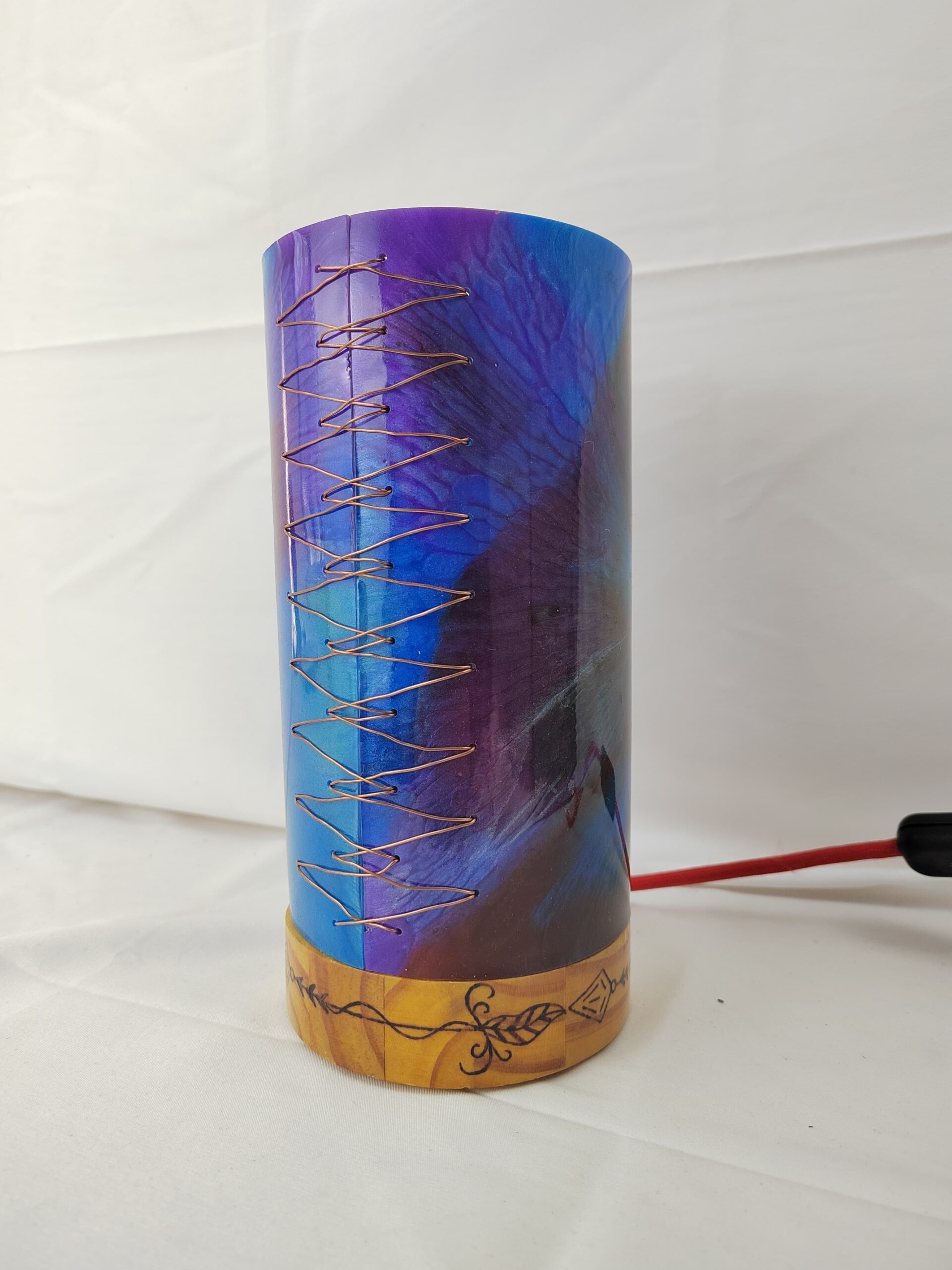 Lámpada artesanal con corpo cilíndrico de resina en cores vibrantes e base de madeira gravada, iluminada desde o interior e unida cunha costura decorativa de fío metálico.