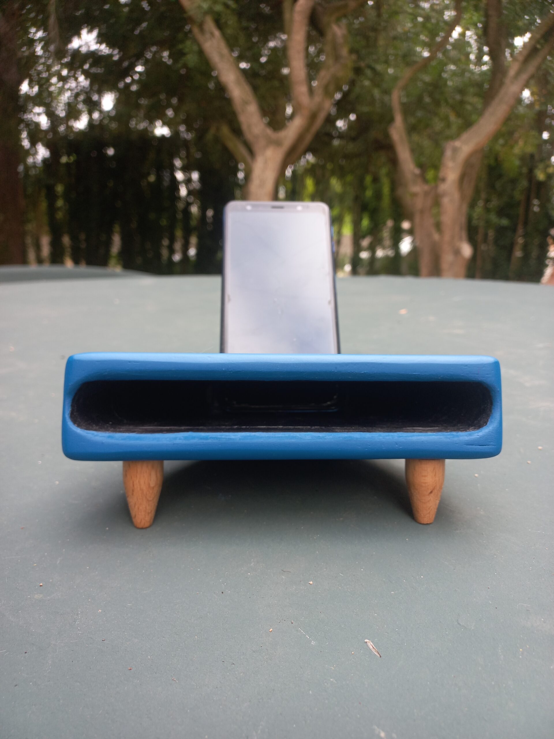 Altofalante pasivo artesanal feito en madeira e resina azul, con cavidade frontal para amplificar o son dun móbil apoiado na parte traseira.