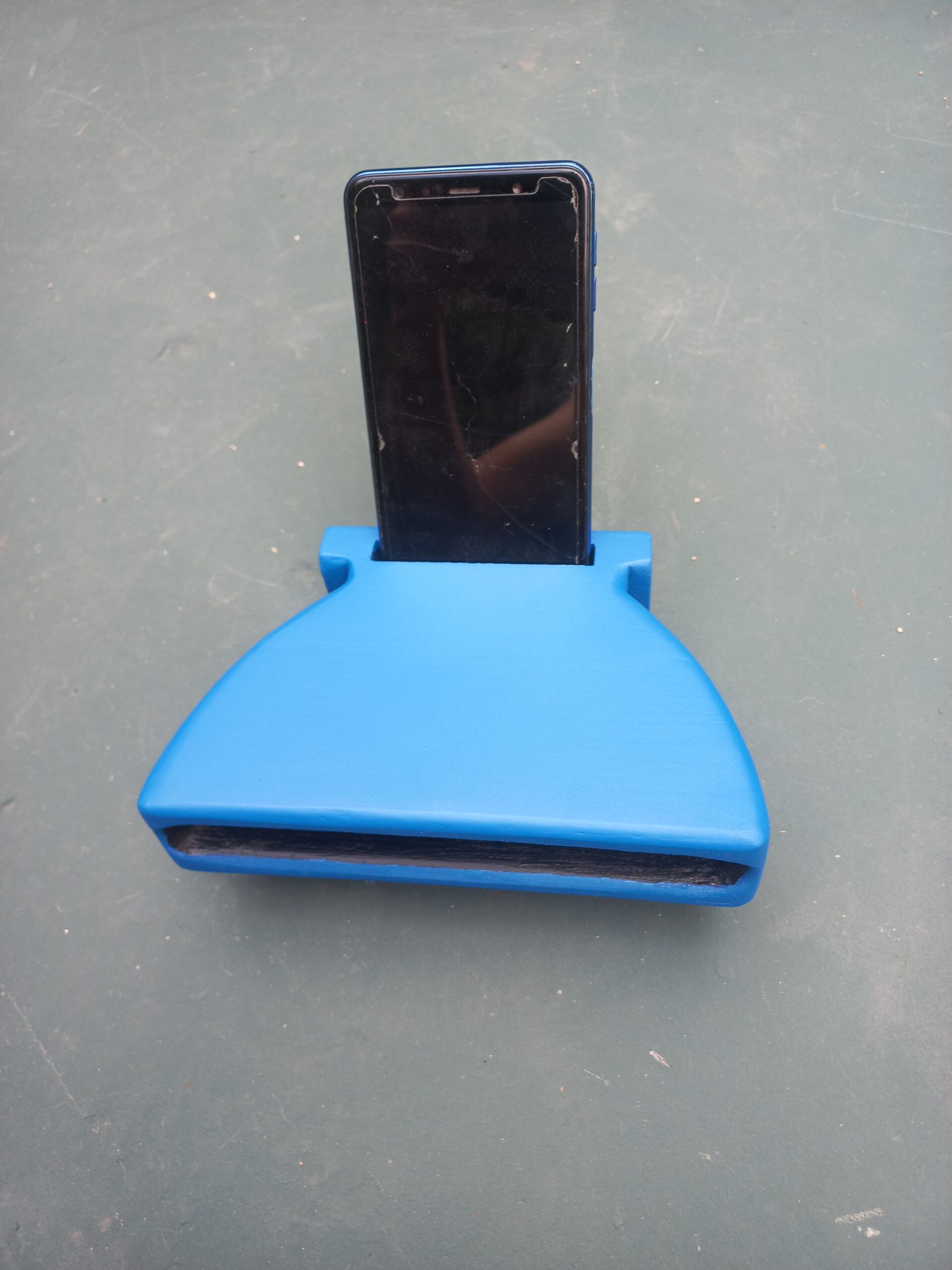 Altofalante pasivo artesanal feito en madeira e resina azul, con cavidade frontal para amplificar o son dun móbil apoiado na parte traseira.