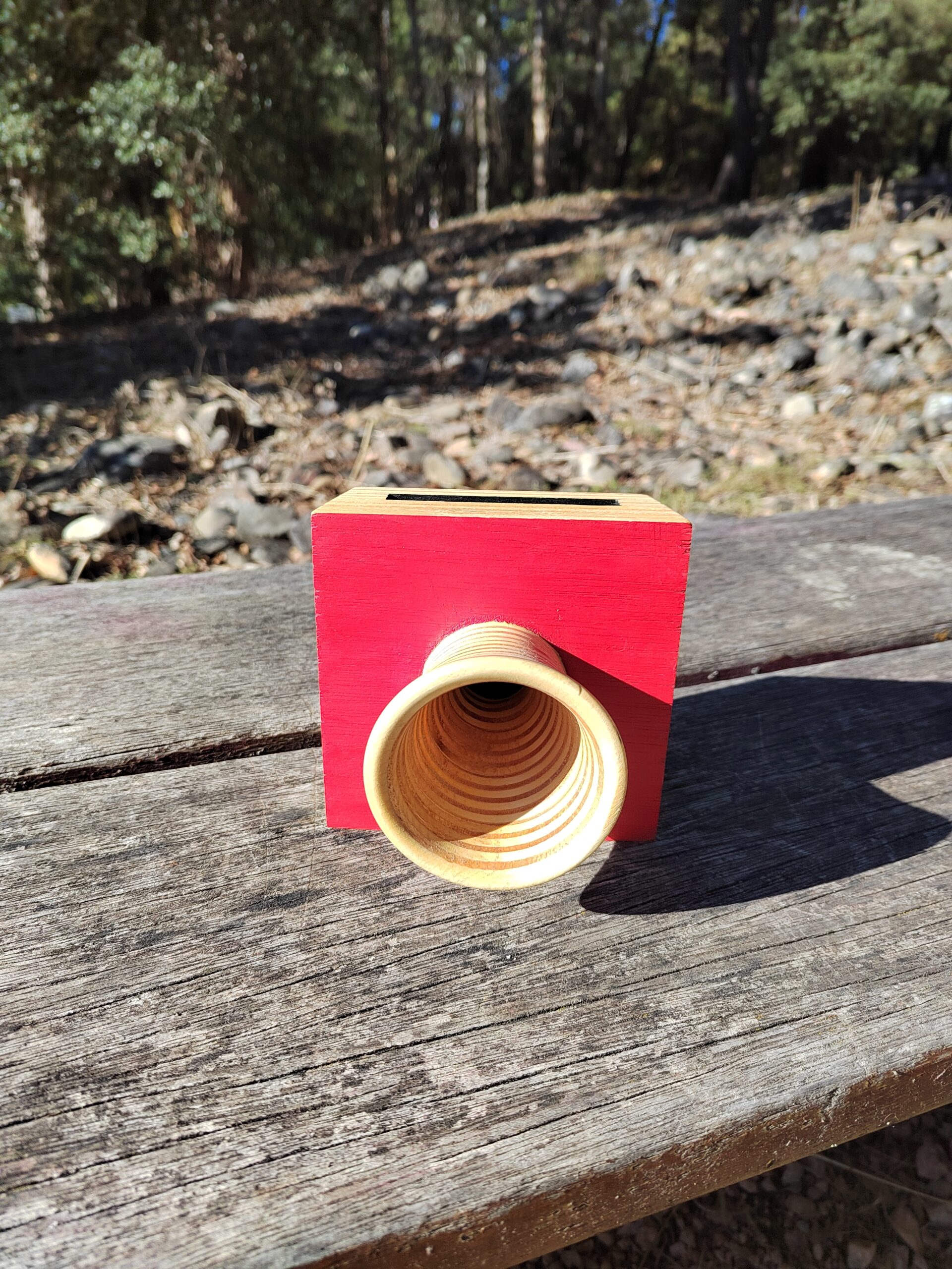 Amplificador de son pasivo artesanal en madeira con panel vermello e bocina frontal torneada, cun smartphone colocado na ranura superior.