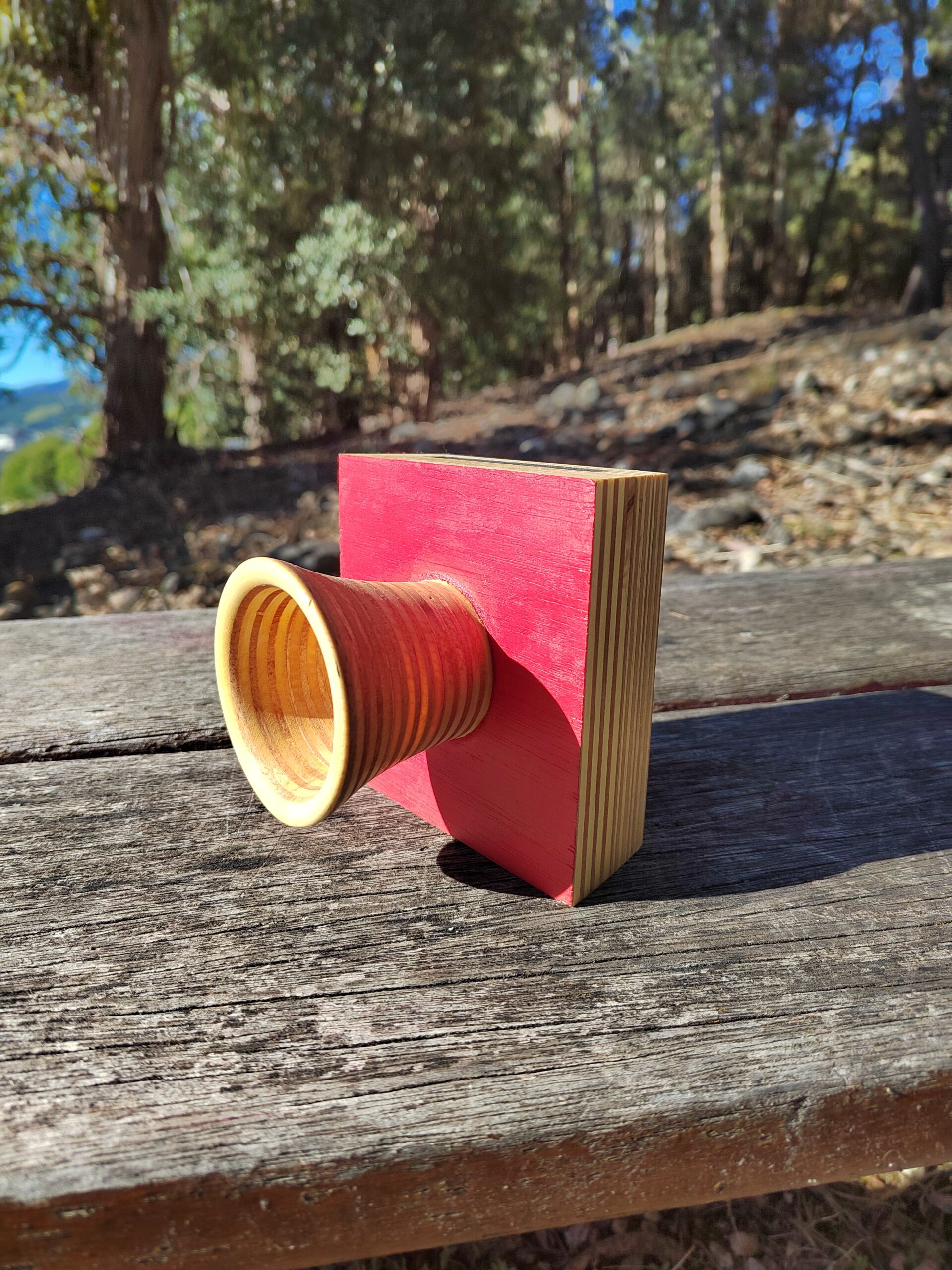 Amplificador de son pasivo artesanal en madeira con panel vermello e bocina frontal torneada, cun smartphone colocado na ranura superior.