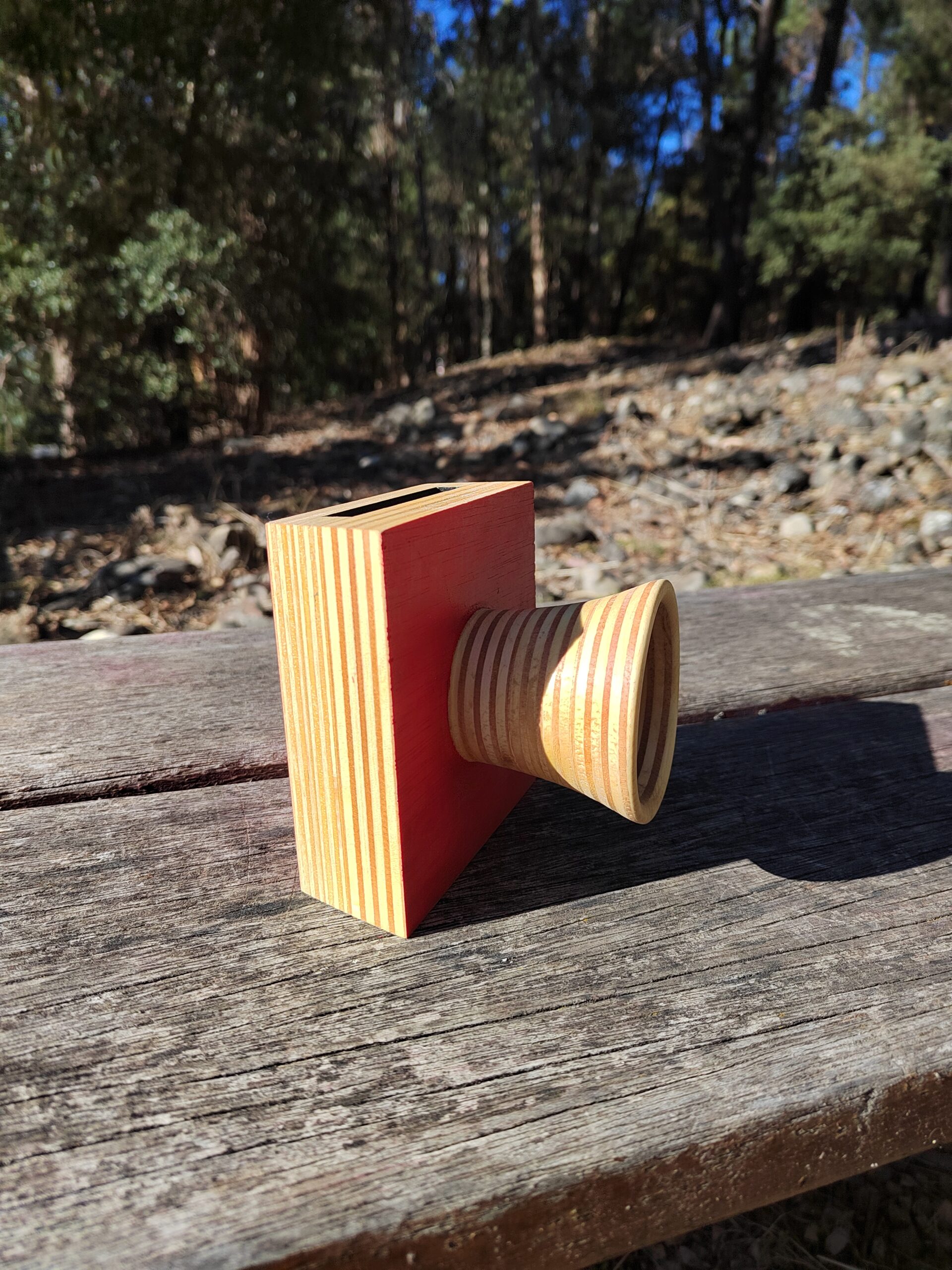 Amplificador de son pasivo artesanal en madeira con panel vermello e bocina frontal torneada, cun smartphone colocado na ranura superior.