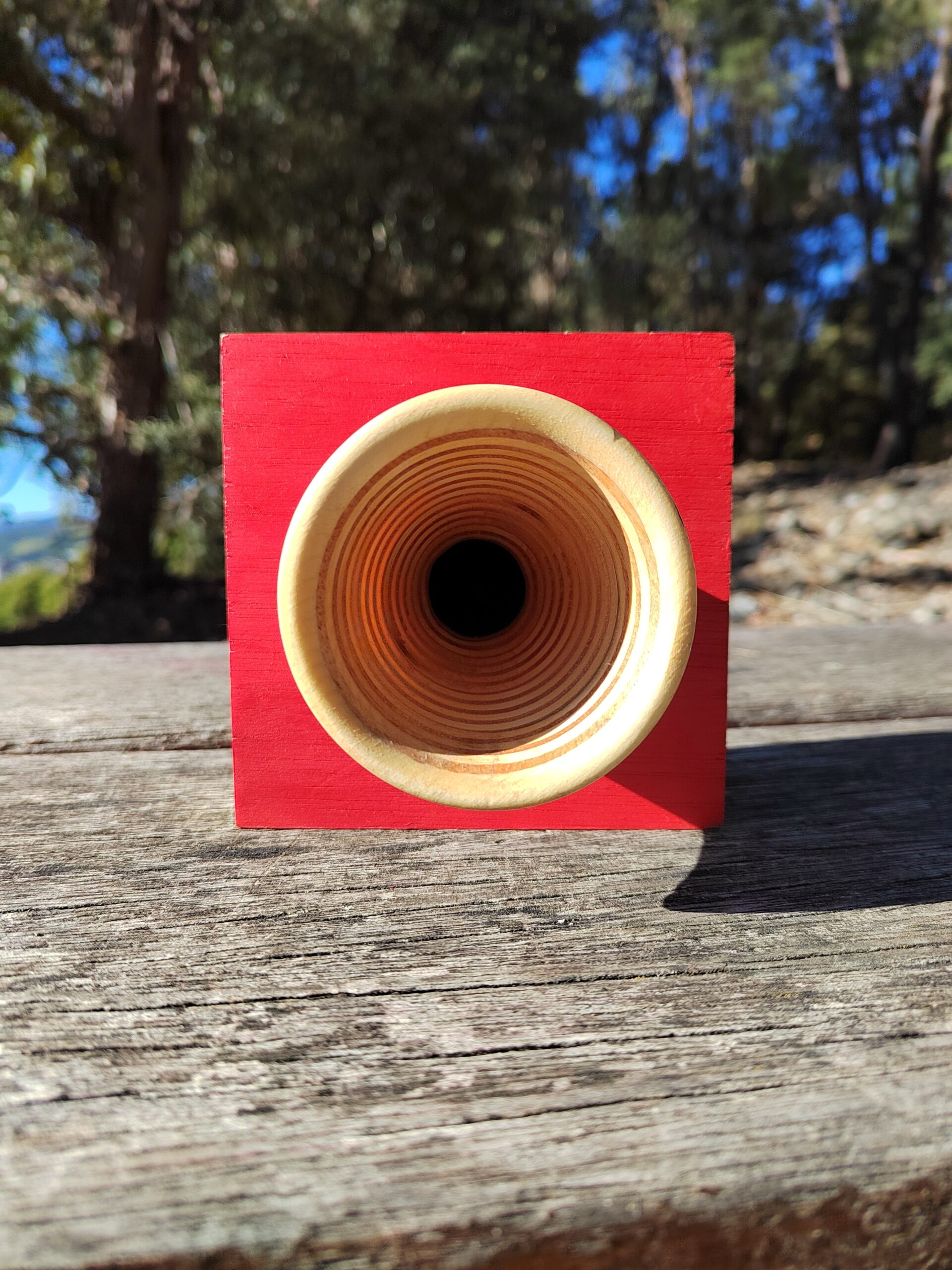 Amplificador de son pasivo artesanal en madeira con panel vermello e bocina frontal torneada, cun smartphone colocado na ranura superior.