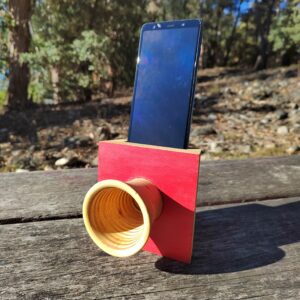 Amplificador de son pasivo artesanal en madeira con panel vermello e bocina frontal torneada, cun smartphone colocado na ranura superior.