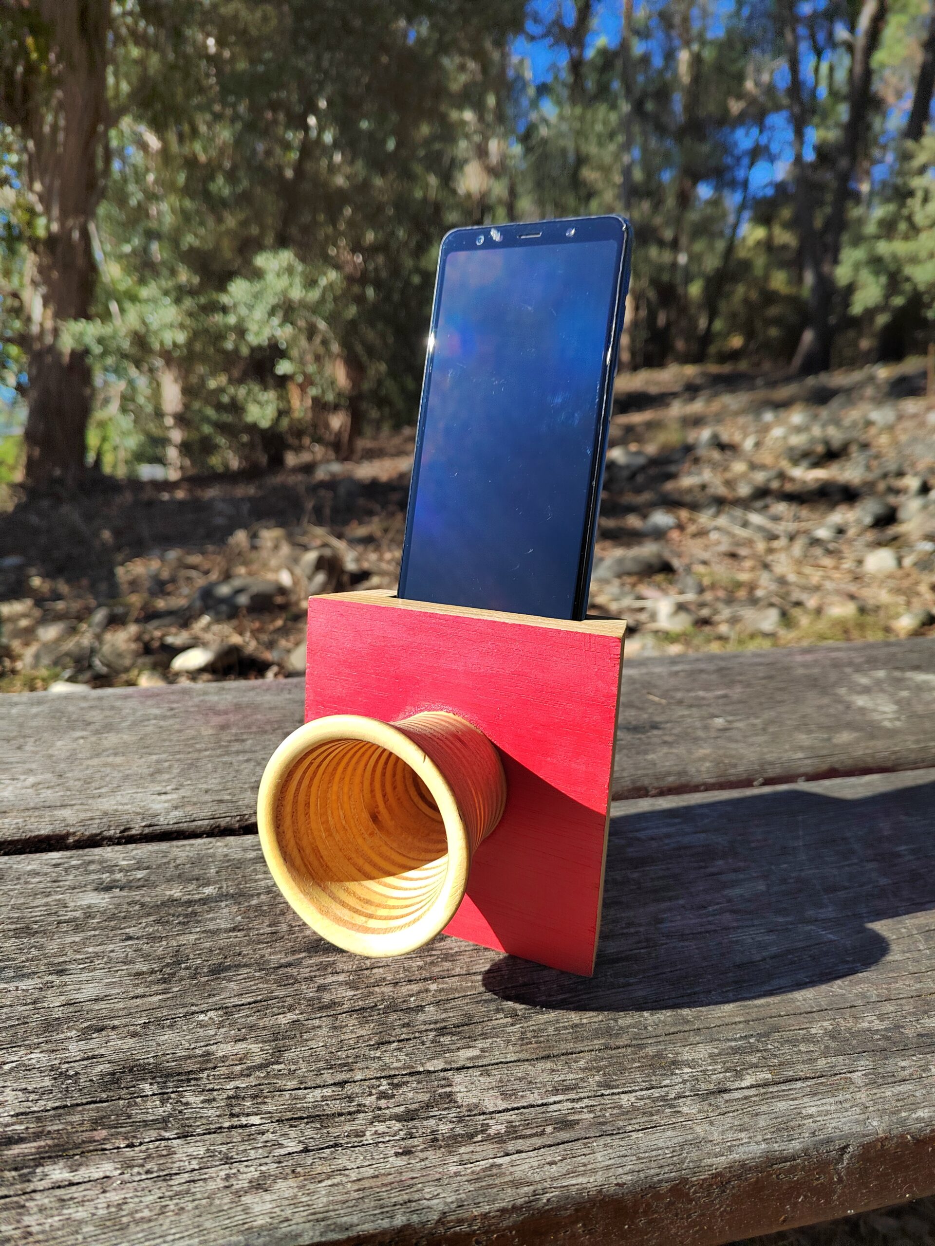 Amplificador de son pasivo artesanal en madeira con panel vermello e bocina frontal torneada, cun smartphone colocado na ranura superior.