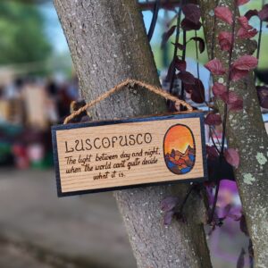 Cartel de madeira pirogravado colgado dunha árbore, coa palabra 'Luscofusco' e o texto 'A luz entre o día e a noite, cando o mundo non sabe ben que é', xunto a unha ilustración ovalada dun solpor sobre montañas.