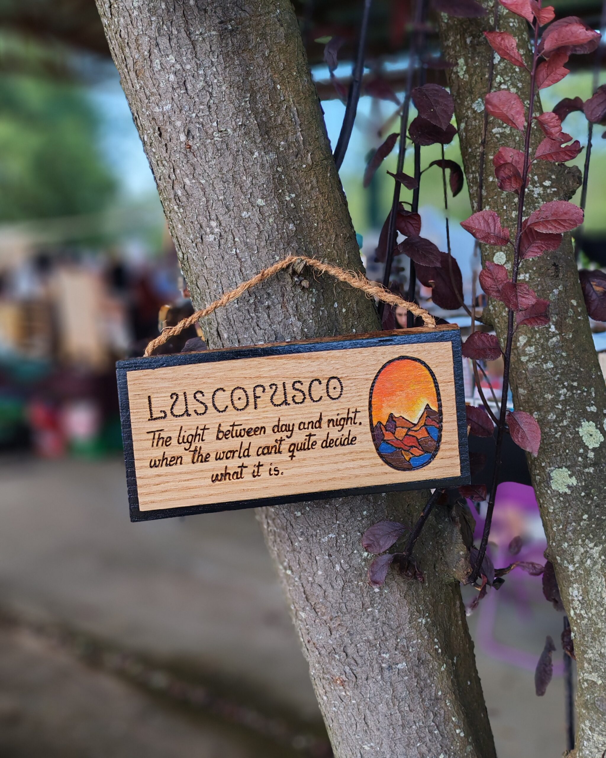 Cartel de madeira pirogravado colgado dunha árbore, coa palabra 'Luscofusco' e o texto 'A luz entre o día e a noite, cando o mundo non sabe ben que é', xunto a unha ilustración ovalada dun solpor sobre montañas.