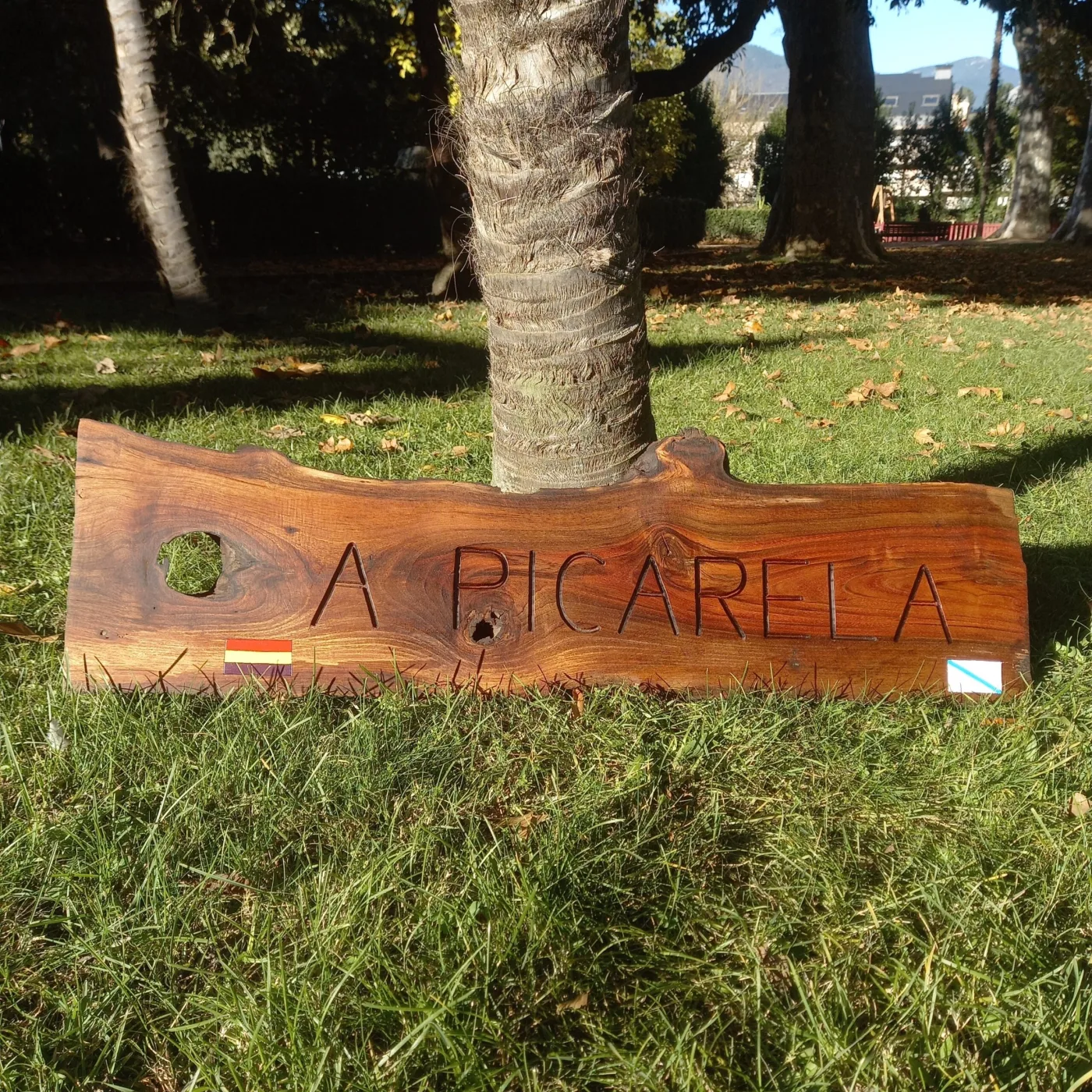 Cartel de madeira A Picarela con bandeiras, apoiado na herba ao pé dunha árbore