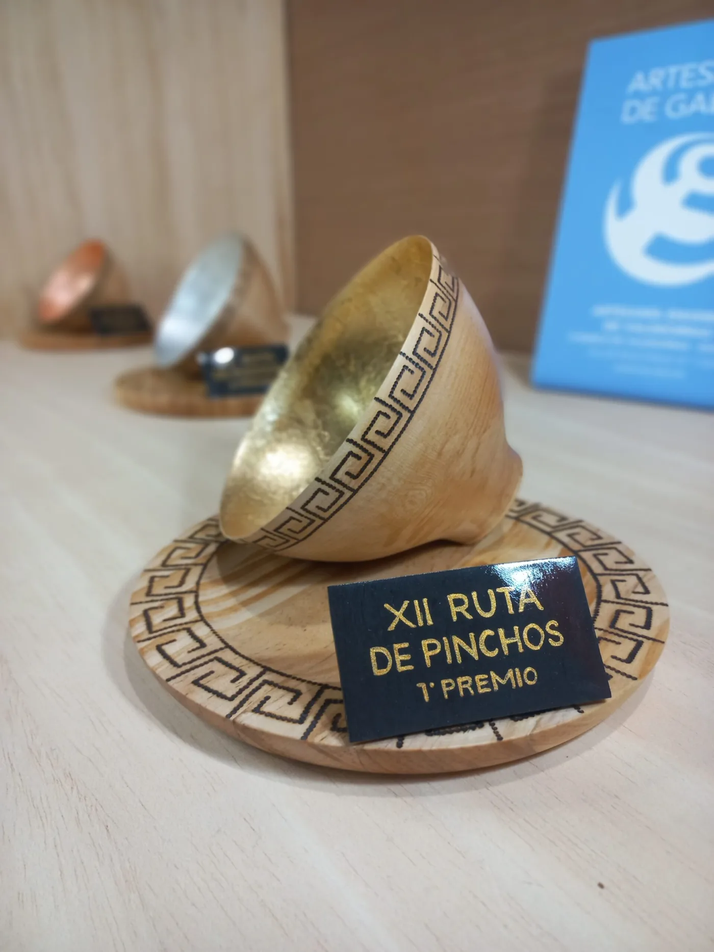 Cunca de madeira con interior dourado sobre un pousa-pratos pirogravado con greca, premio da XII Ruta de Pinchos