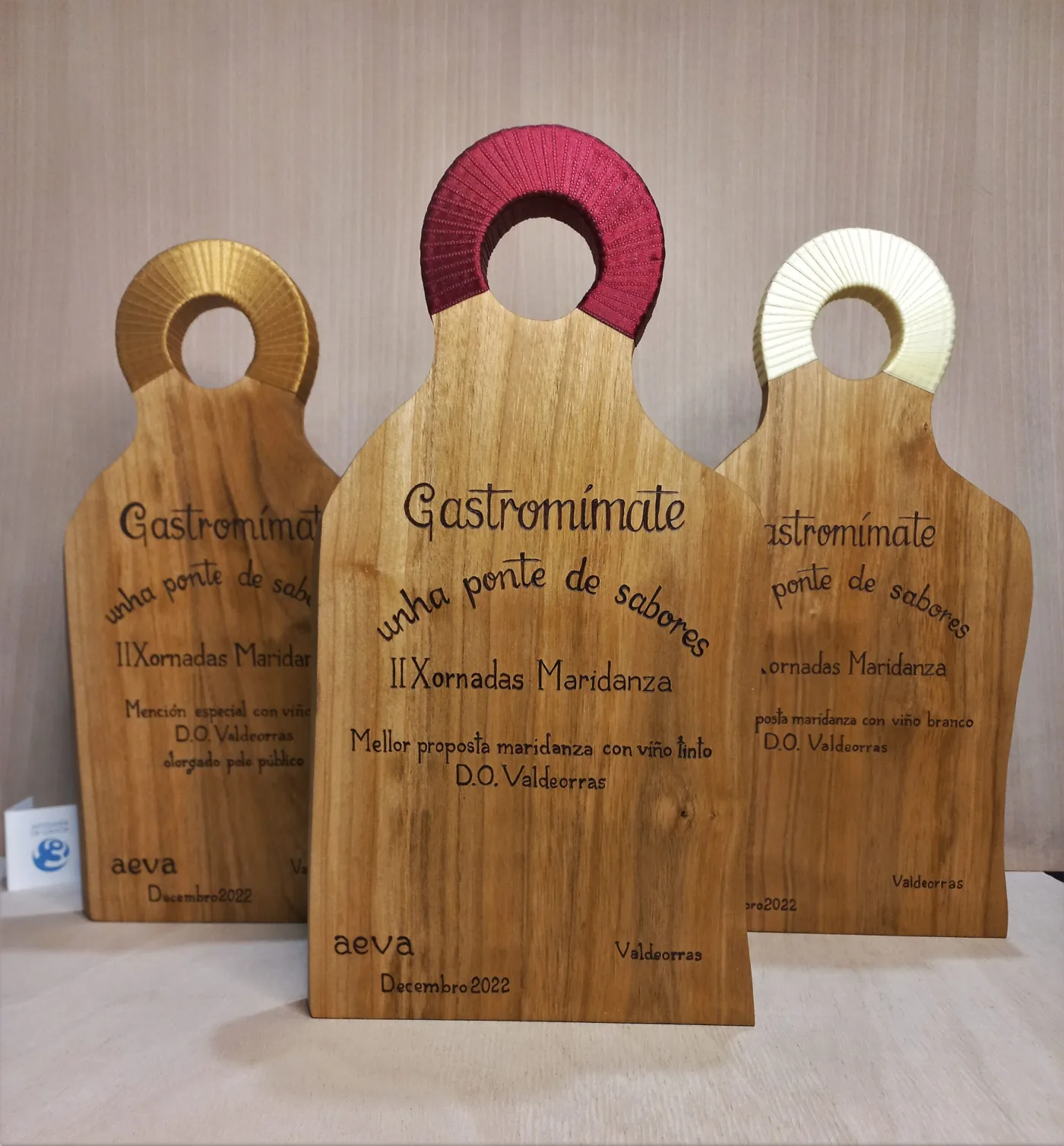 Tres galardóns de madeira con asa de cores e texto pirogravado para as Xornadas Gastromimate de Valdeorras