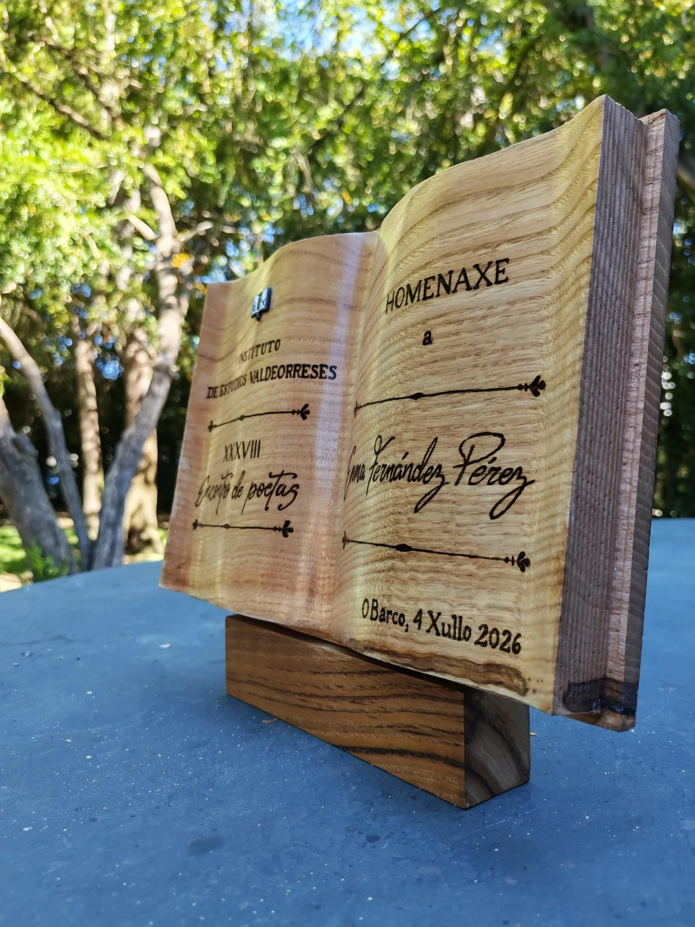 Libro de madeira aberto con texto pirogravado de homenaxe, sobre unha peana de madeira, ao aire libre