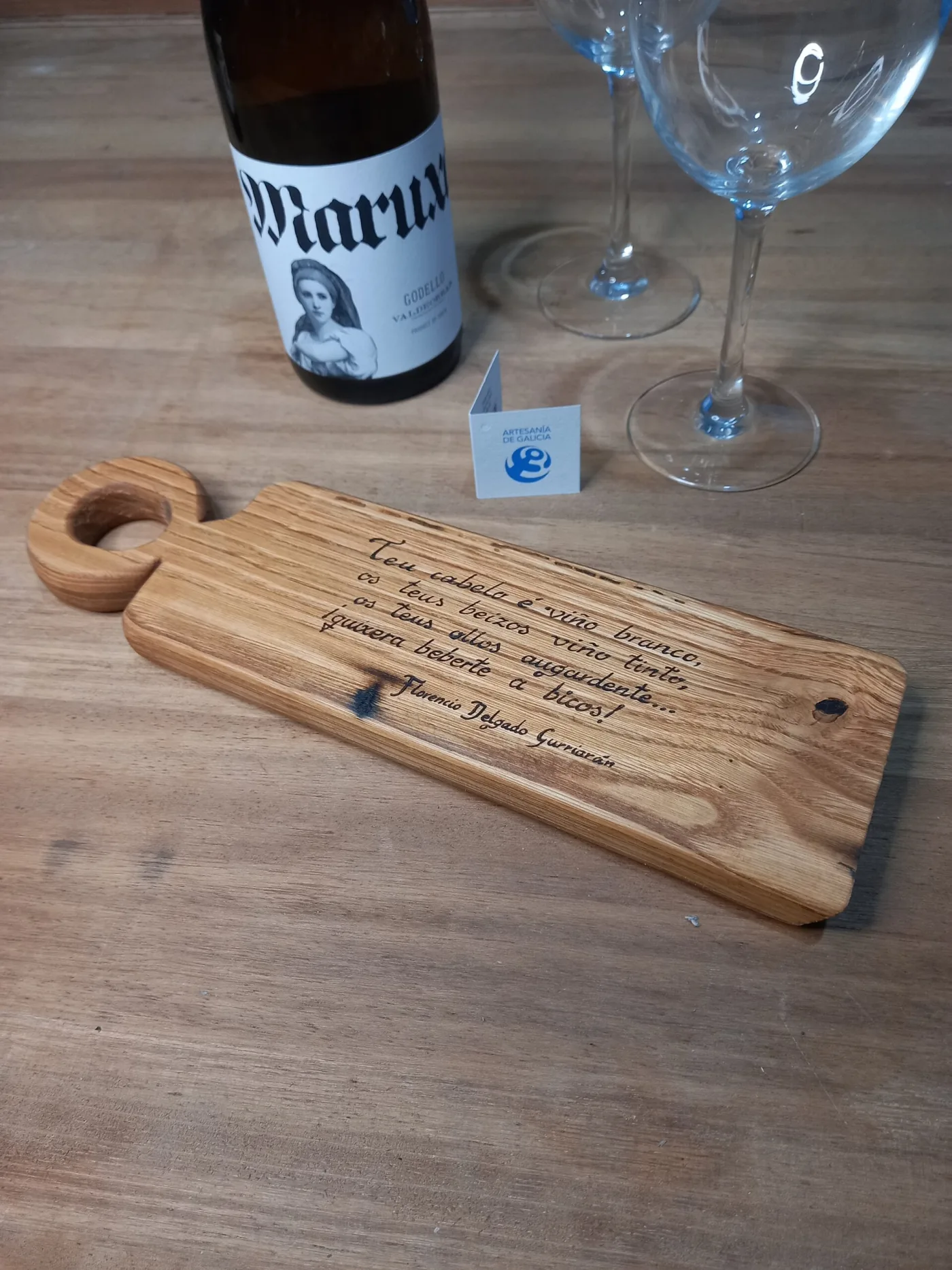 Táboa de madeira con asa e dedicatoria pirogravada, xunto a unha botella de viño e dúas copas