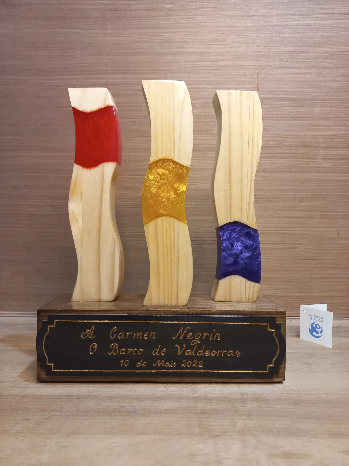 Trofeo de homenaxe con tres tiras onduladas de madeira e resina vermella, amarela e violeta sobre unha peana con placa